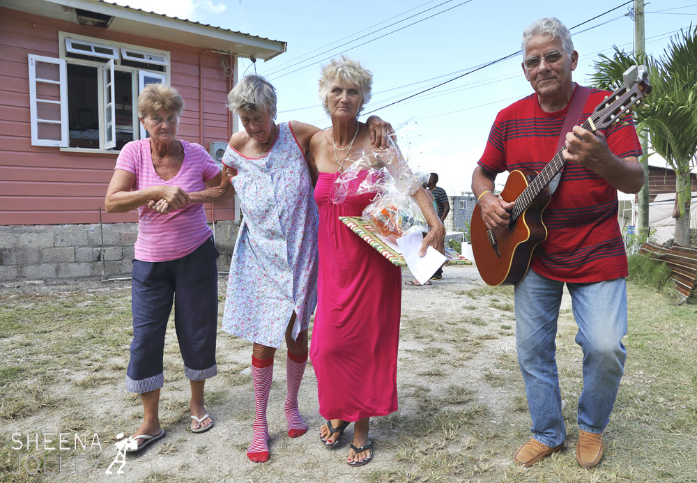 Erlene Downie being helped back to her own house by daughters Hazel (on left) and Ann (on right) and serenaded by Willie Kerr of The Merrymen band so well known in Barbados for years. Ann is holding the hamper of food and Christmas present given to Erlene on Love Day by Terry Arthur. Terry started the Love Day charity 5 years ago visiting vagrants, homeless and sick but this is their first visit to the "poor white" community on the east coast.