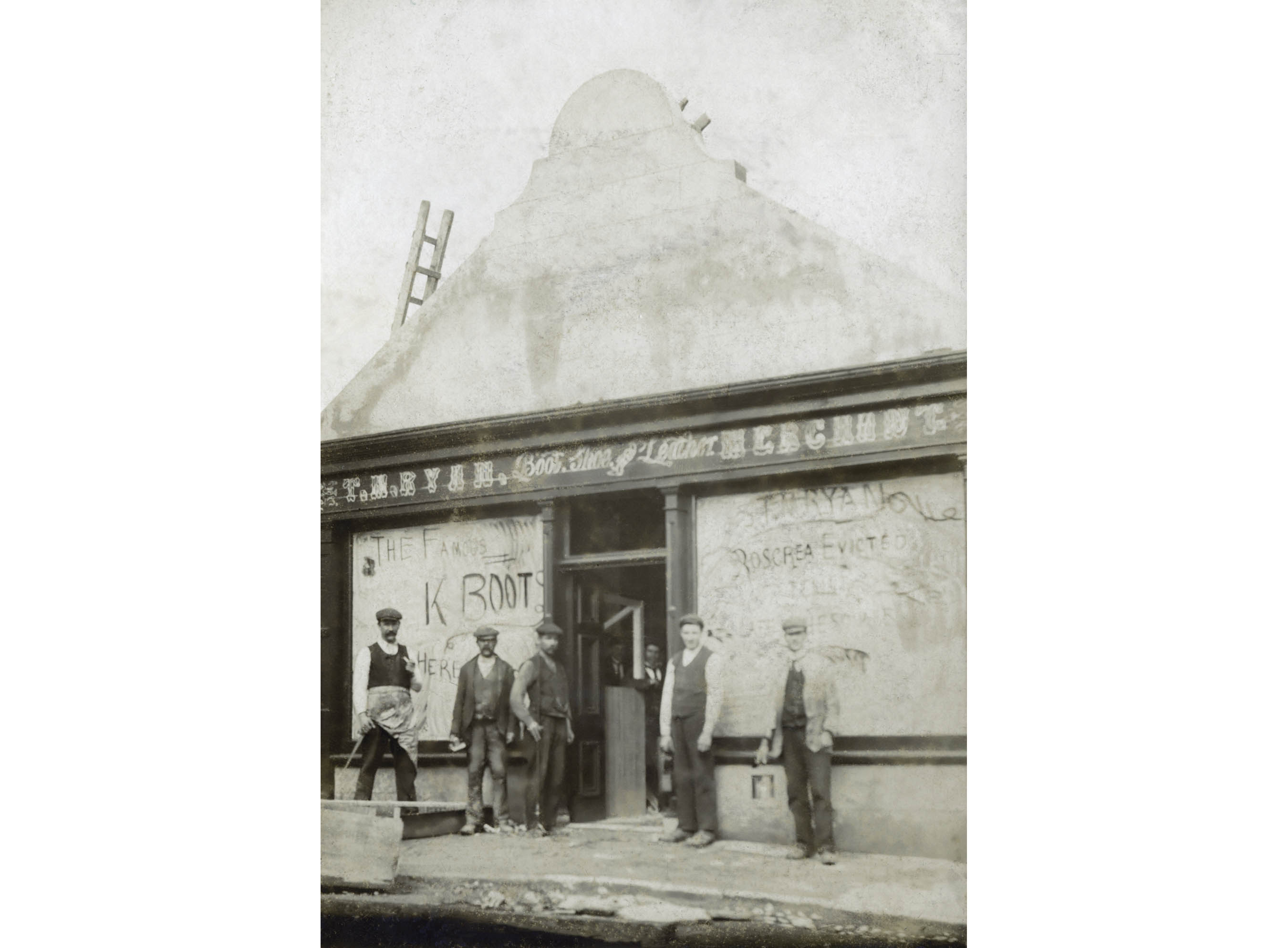 Men stand in front of a shoe and leather merchant store with boarded up windows. Writing on the right window reads “T.M.Ryan Roscrea Evicted.” Roscrea, Co. Tipperary, c. 1888.