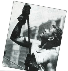 A black and white photo of an Irish constructor worker holds a guide rope as the great beam goes up on the Empire State Building. 