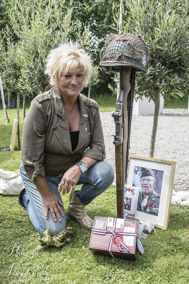 Ellen Womer, daughter of Sgt. Jack Womer, kneels beside the olive tree she planted with her father in his honor in 2013. The D-Day veteran died before the 2014 commemoration. Ellen brought her father's photo and ashes to the ceremony. The upturned Garand rifle with fixed bayonet and helmet is a tribute Ashe puts beside the tree of each hero who died in the preceding year. Jack Womer’s ashes were permanently interred elsewhere in Normandy a few days later.